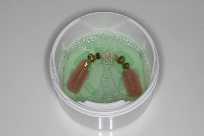 Die Reinigung erfolgt über 10-15 Minuten. Die Reinigungslösung verliert mit der Zeit an Wirkung und spätestens nach 30 Minuten sollte die Reinigungsflüssigkeit abgekippt und die Prothese nochmals mit klarem Wasser abgespült werden.